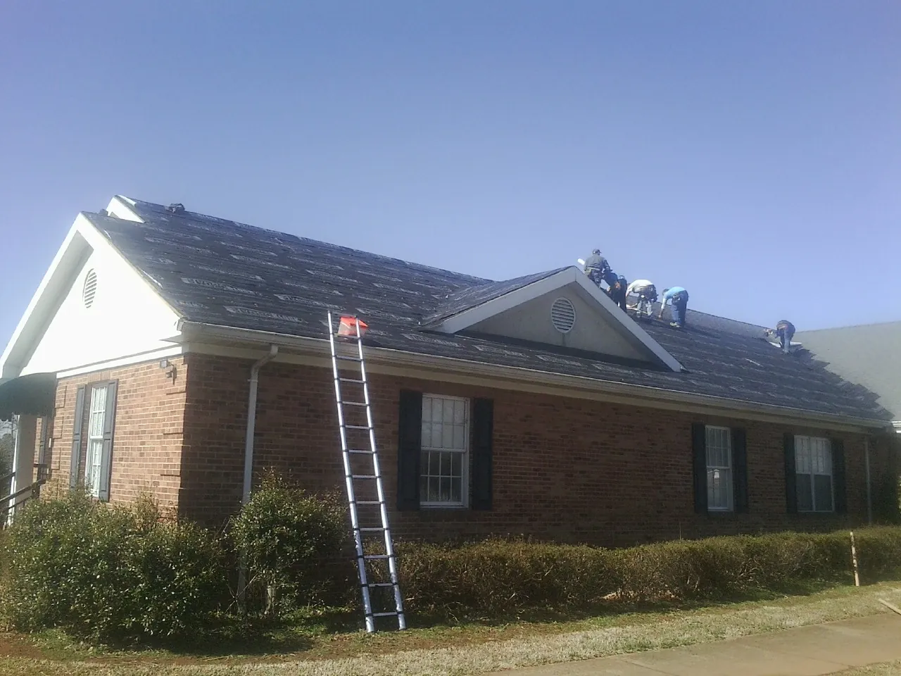 Crew tearing shingles off a brick ranch roof with a ladder set — Metal Roof Repair work by our crew in Rices Landing