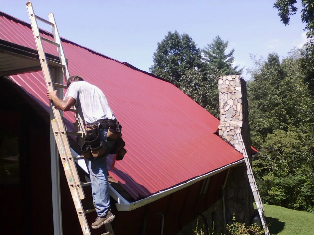 Roofer on a ladder beside a red metal roof and stone chimney in Rices Landing
